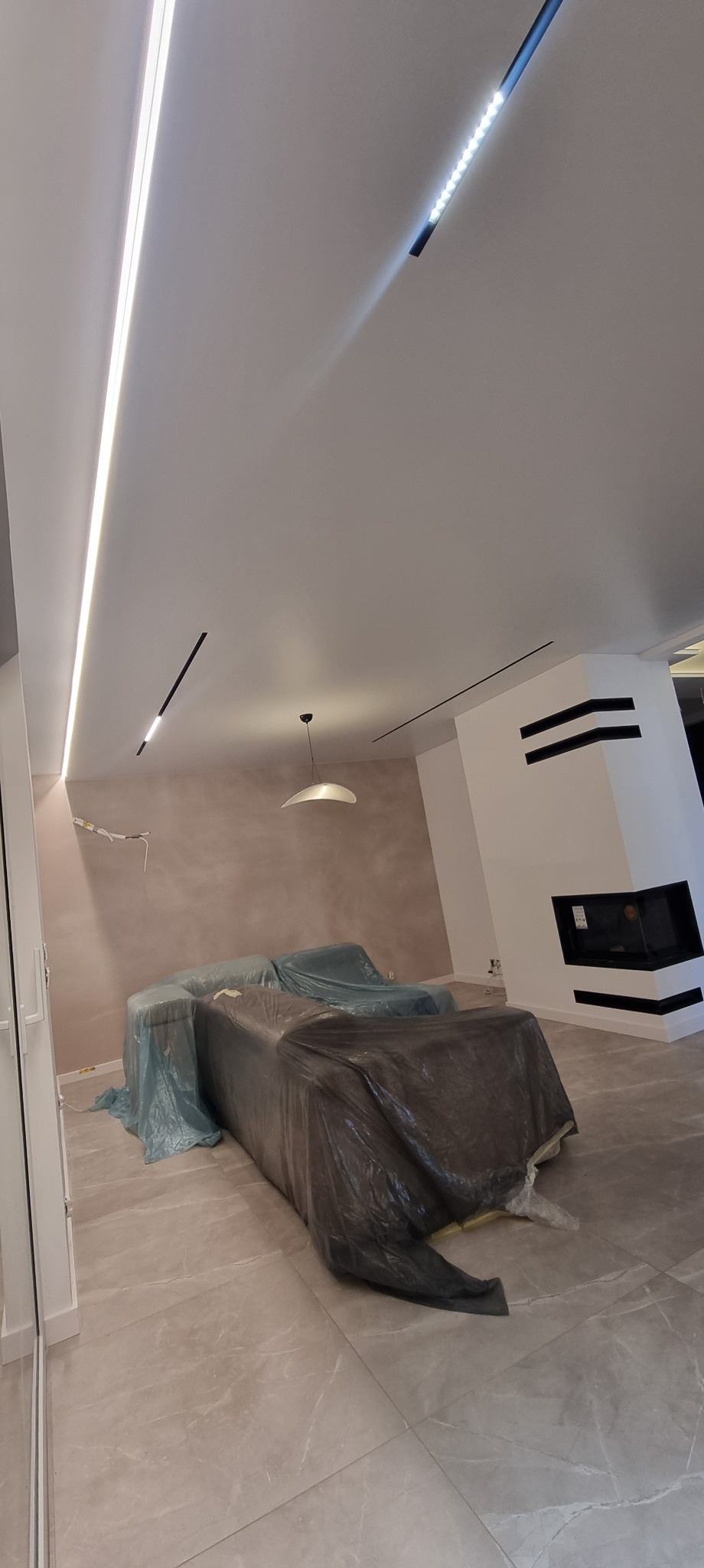Nowoczesne oświetlenie sufitowe LED w salonie z kominkiem, meble zabezpieczone folią ochronną. Widoczne listwy LED i lampa wisząca.