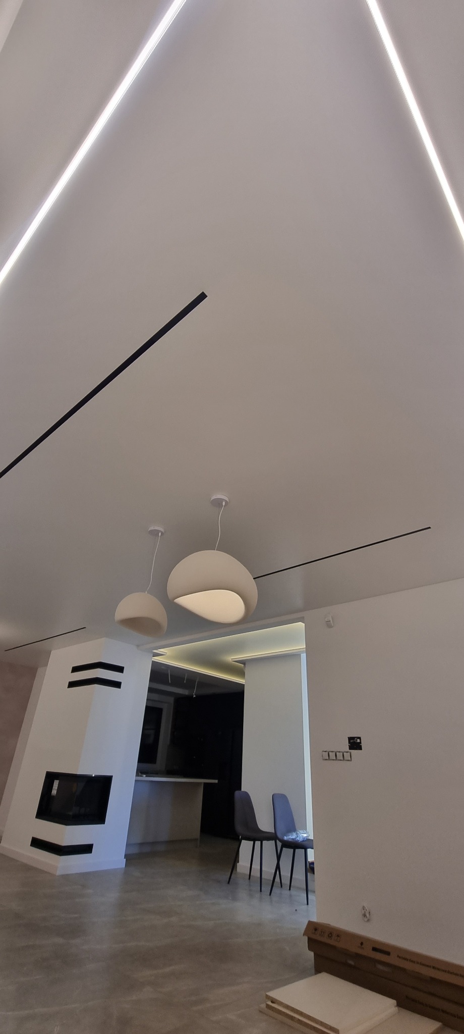 Nowoczesny salon z oświetleniem LED w suficie, lampami wiszącymi i minimalistycznym kominkiem. Widoczne krzesła i elementy wykończenia wnętrz.