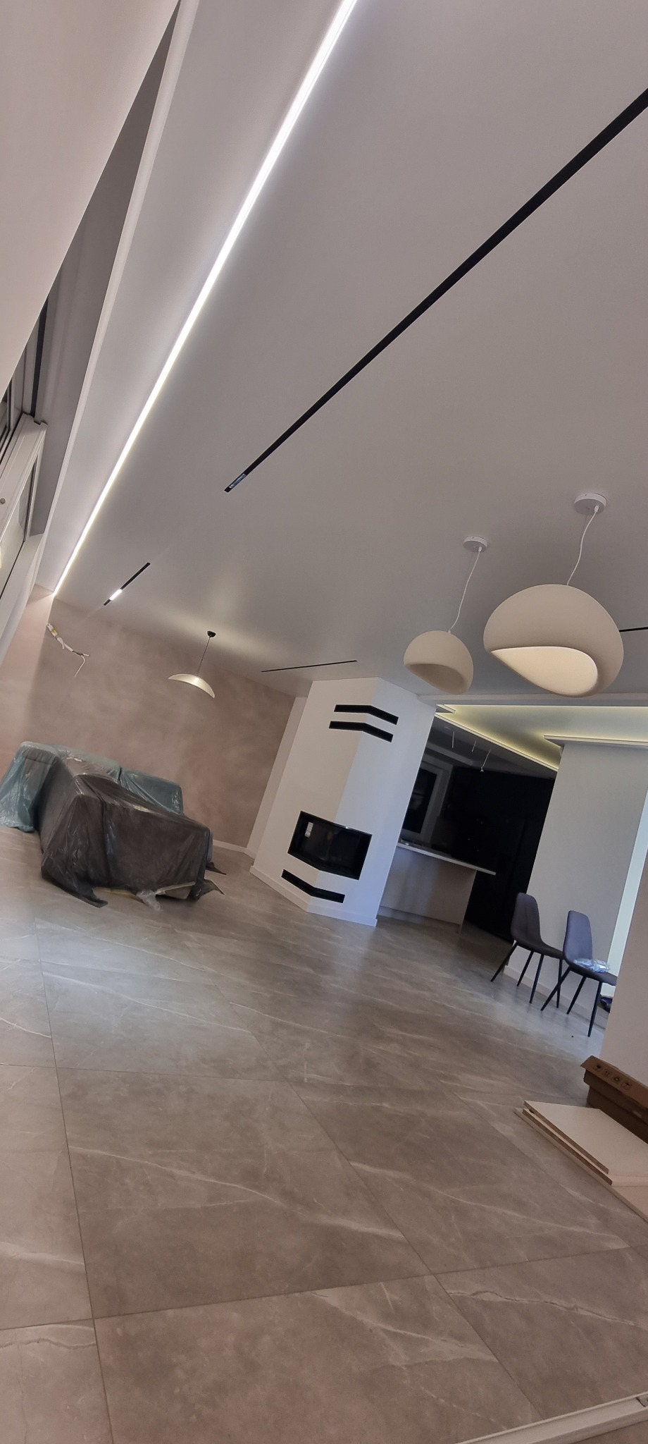 Nowoczesne oświetlenie sufitowe w przestronnym salonie z kominkiem i szarymi płytkami podłogowymi. Widoczne lampy wiszące i listwy LED.