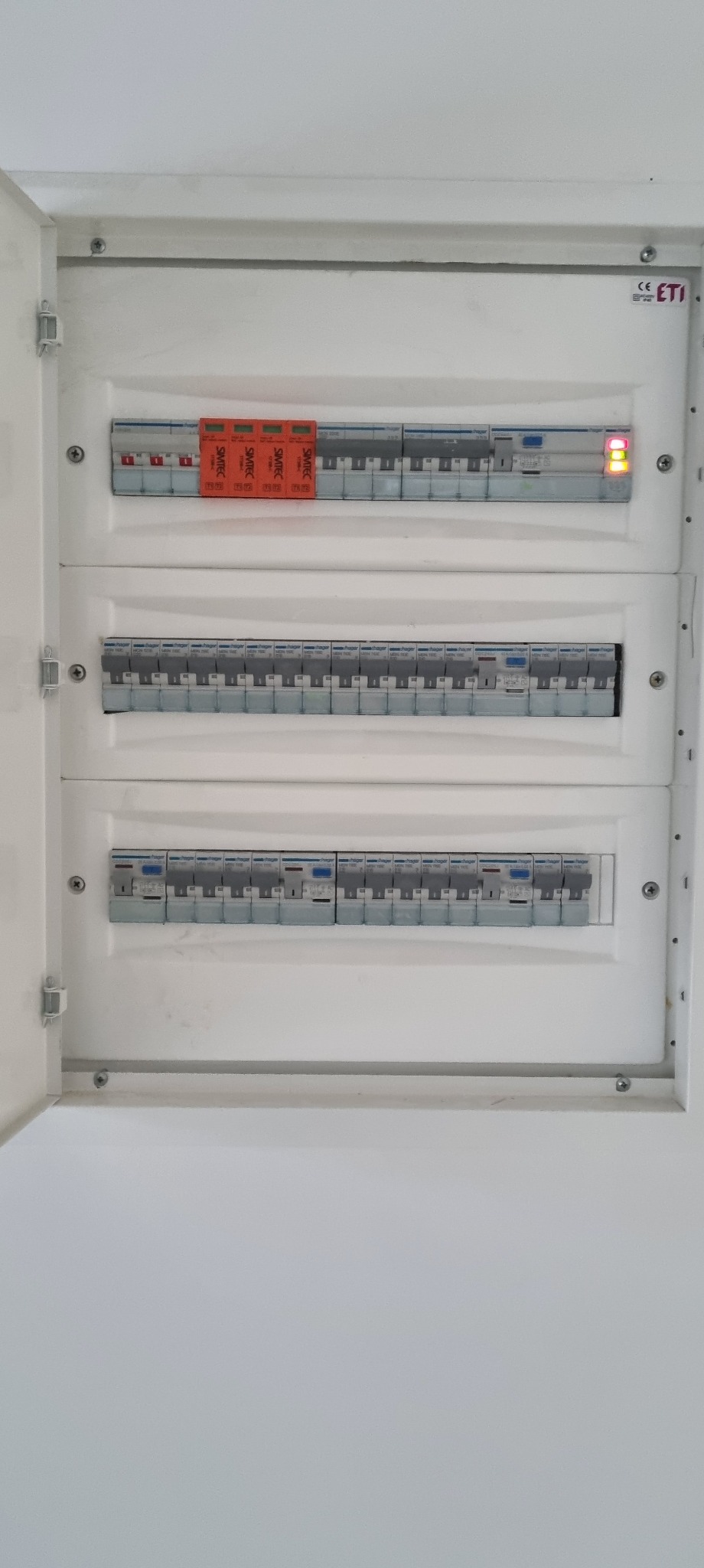 Otwarta, biała skrzynka rozdzielcza z trzema rzędami bezpieczników. Na górze czerwone ograniczniki przepięć. Dwie diody sygnalizacyjne świecą na pomarańczowo.