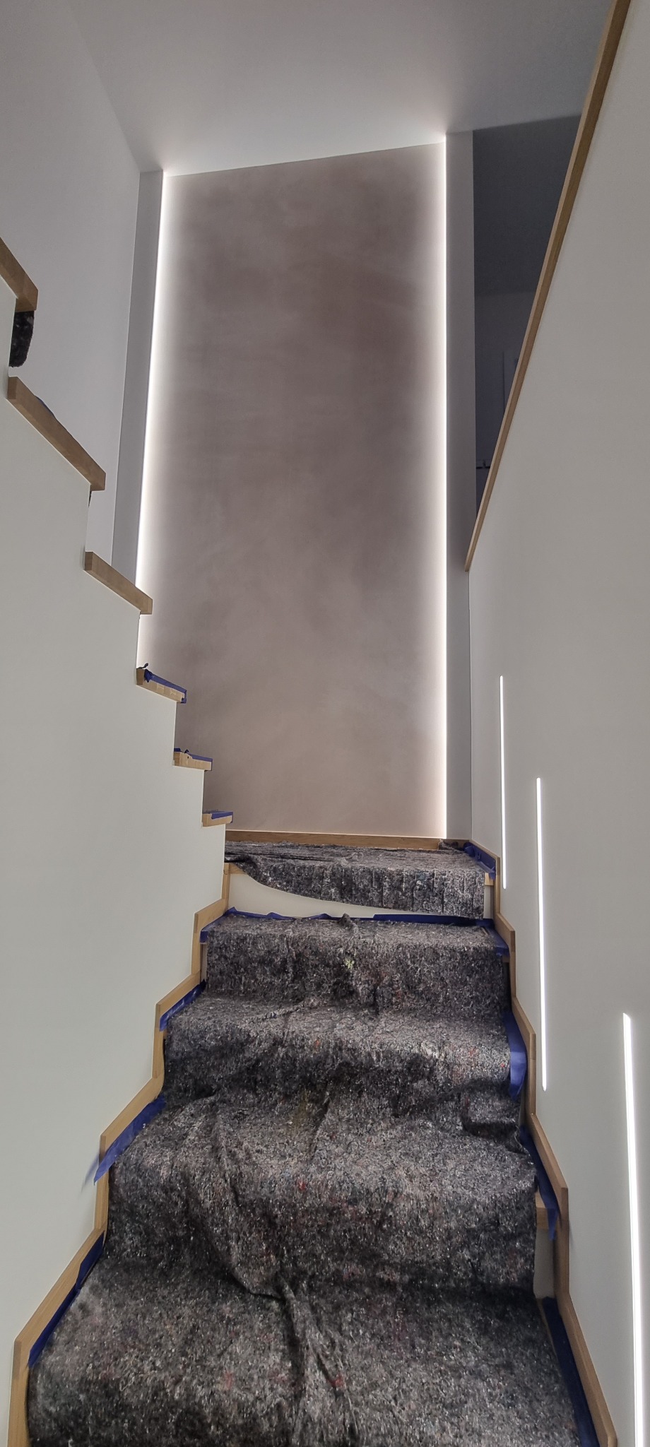 Nowoczesne schody z podświetleniem LED wzdłuż ściany i stopni, zabezpieczone folią malarską. Minimalistyczny design, jasne kolory, subtelne oświetlenie.