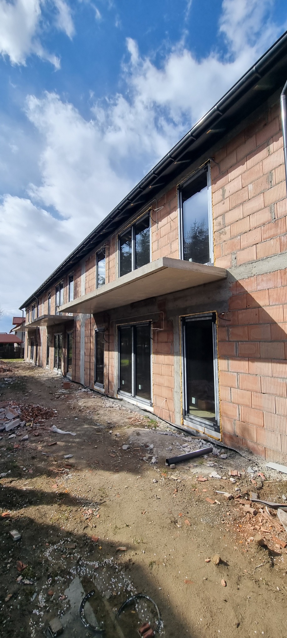 Budynek w trakcie budowy z widocznymi oknami i balkonami, wykonany z czerwonej cegły. Widoczne elementy instalacji na elewacji. Ujęcie z dołu, w tle błękitne niebo.