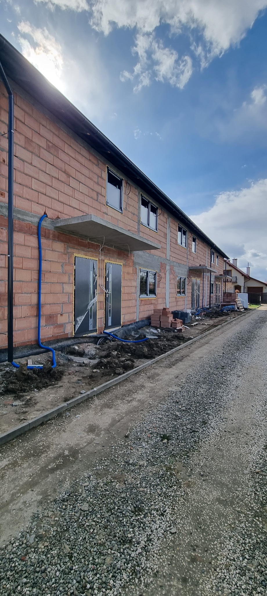 Surowy stan budynku w trakcie budowy: widoczne cegły, okna z czarnymi ramami, instalacja rur na zewnątrz. Ujęcie z perspektywy drogi szutrowej.