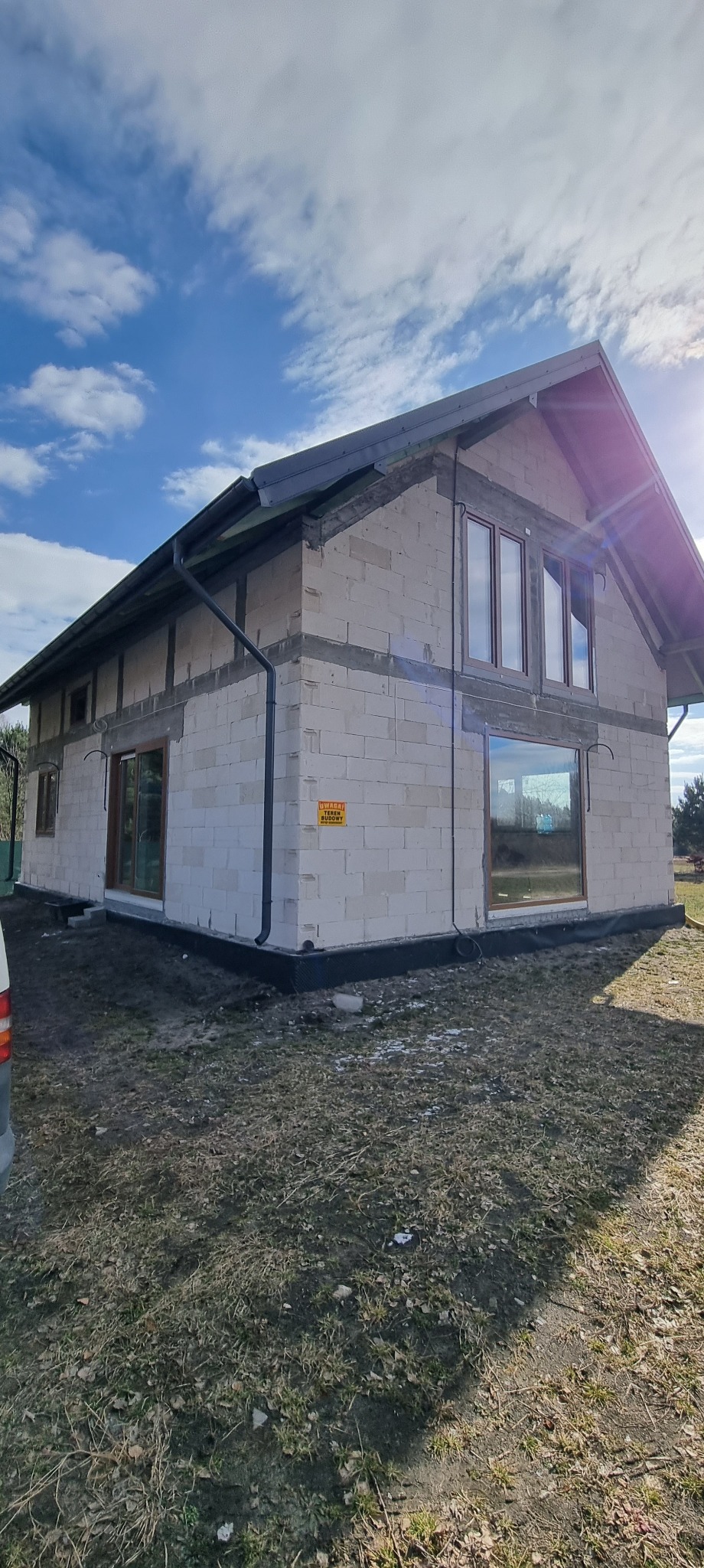 Surowy stan domu jednorodzinnego z białego bloczka, z zamontowanymi oknami i orynnowaniem, na tle błękitnego nieba z chmurami. Widoczny fragment samochodu.