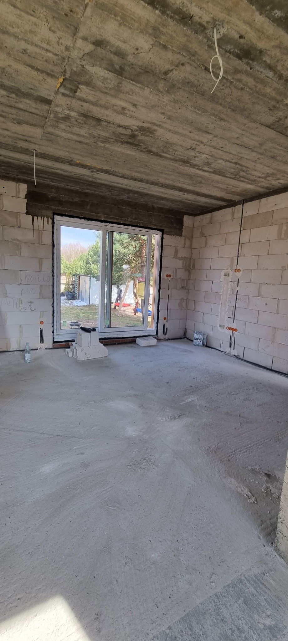 Surowy stan wnętrza: ściany z betonu komórkowego, okno z widokiem na zewnątrz, instalacja elektryczna w bruzdach. W tle butelka wody i worki z zaprawą.