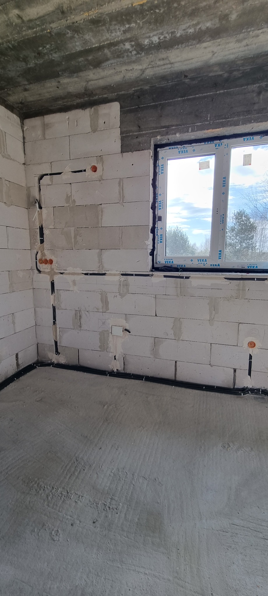 Surowy stan: Instalacja elektryczna w peszlach na ścianach z betonu komórkowego, widoczne puszki instalacyjne. Okno z widokiem na zewnątrz. Szare ściany i podłoga.