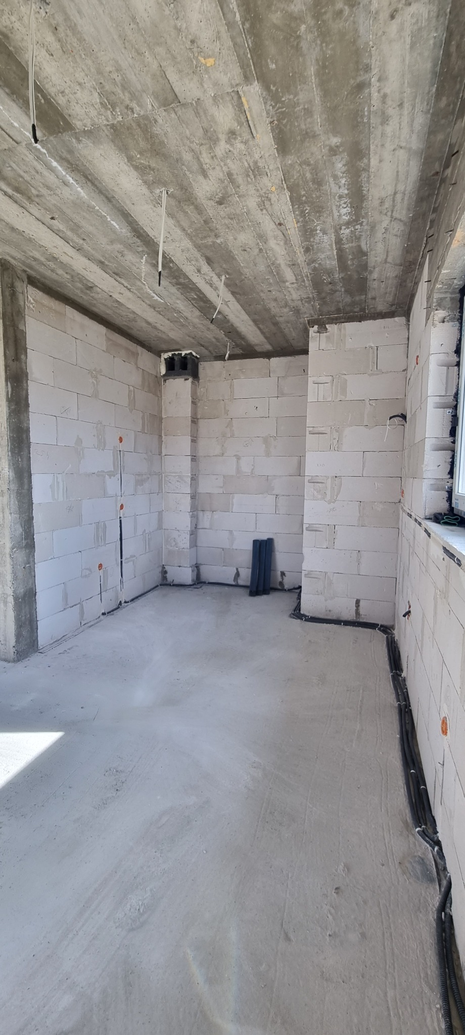 Surowy stan wnętrza z instalacją elektryczną w bruzdach ścian i przewodami zwisającymi z sufitu. Widoczne bloczki betonowe i wylewka podłogowa.