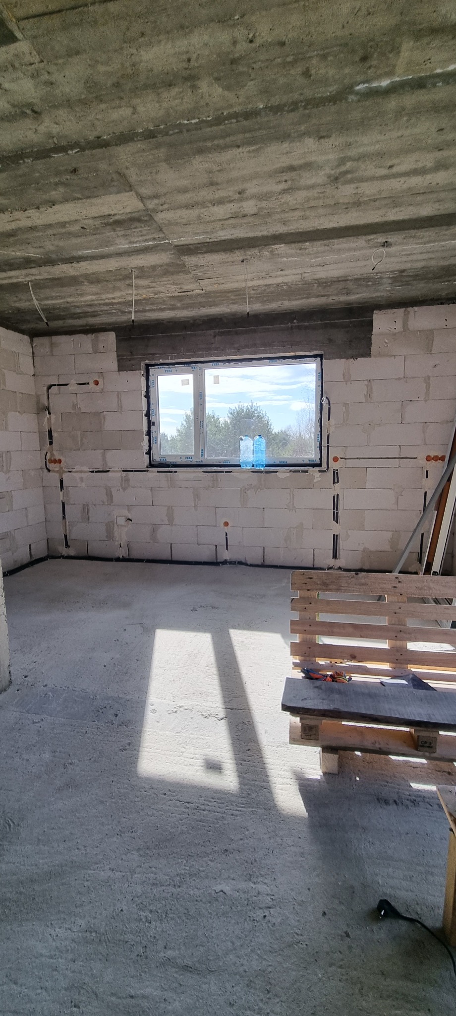 Surowy stan wnętrza z widoczną instalacją elektryczną w bruzdach ścian, przygotowaną do dalszych prac wykończeniowych. Paleta z narzędziami w rogu pomieszczenia.