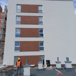 BARTŁOMIEJ GAŹDZIAK USŁUGI BUDOWLANE - Budynek wielorodzinny z elewacją w trakcie wykańczania, widoczne fragmenty ceglanej okładziny wokół okien, robotnik w żółtym kasku i kamizelce odblaskowej na dachu, kominy wentylacyjne, rusztowania...