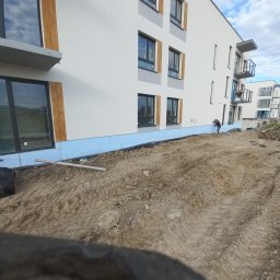 BARTŁOMIEJ GAŹDZIAK USŁUGI BUDOWLANE - Prace przygotowawcze do ocieplenia elewacji budynku wielorodzinnego, widoczna warstwa izolacji przy gruncie i robotnik przy elewacji.