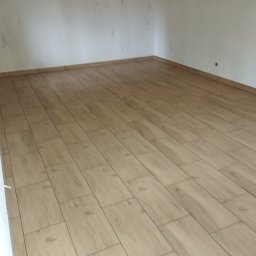 BARTŁOMIEJ GAŹDZIAK USŁUGI BUDOWLANE - Pusta przestrzeń w rogu pokoju z nową, jasną podłogą z paneli imitujących drewno i pomalowanymi na biało ścianami z widocznymi zabrudzeniami.