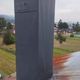 Szary, metalowy komin z geometrycznym wzorem na dachu z blachy trapezowej, w tle krajobraz wiejski z domami i wzgórzami.