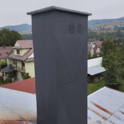 Nowoczesny, grafitowy komin z geometrycznym wzorem na dachu z blachy falistej, w tle zabudowa wiejska i zalesione wzgórza.