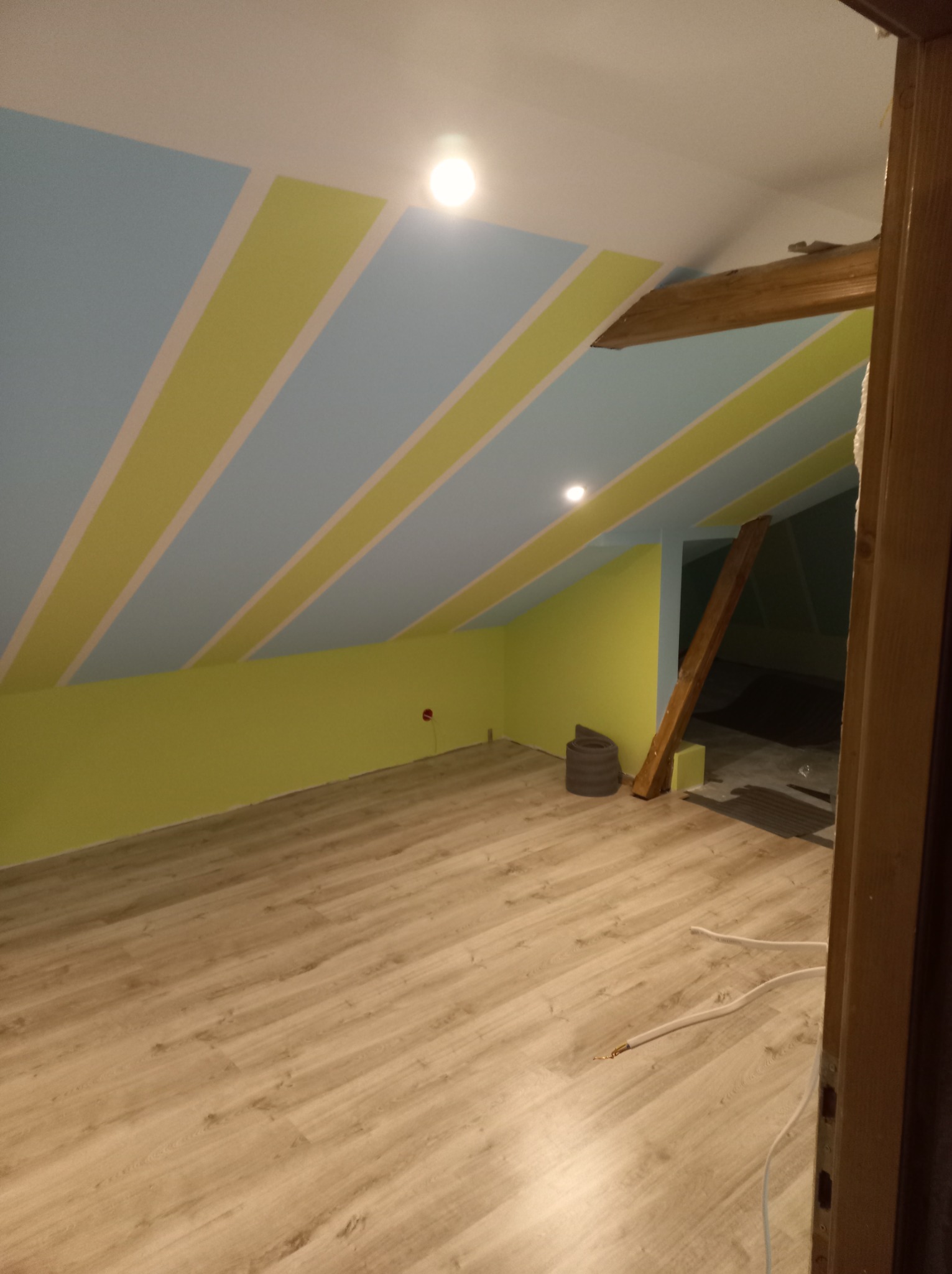 Pomalowane poddasze z drewnianą belką konstrukcyjną,  żółto-niebieskie pasy na skośnym suficie, jasne panele podłogowe, widoczne kable elektryczne i fragment drzwi.
