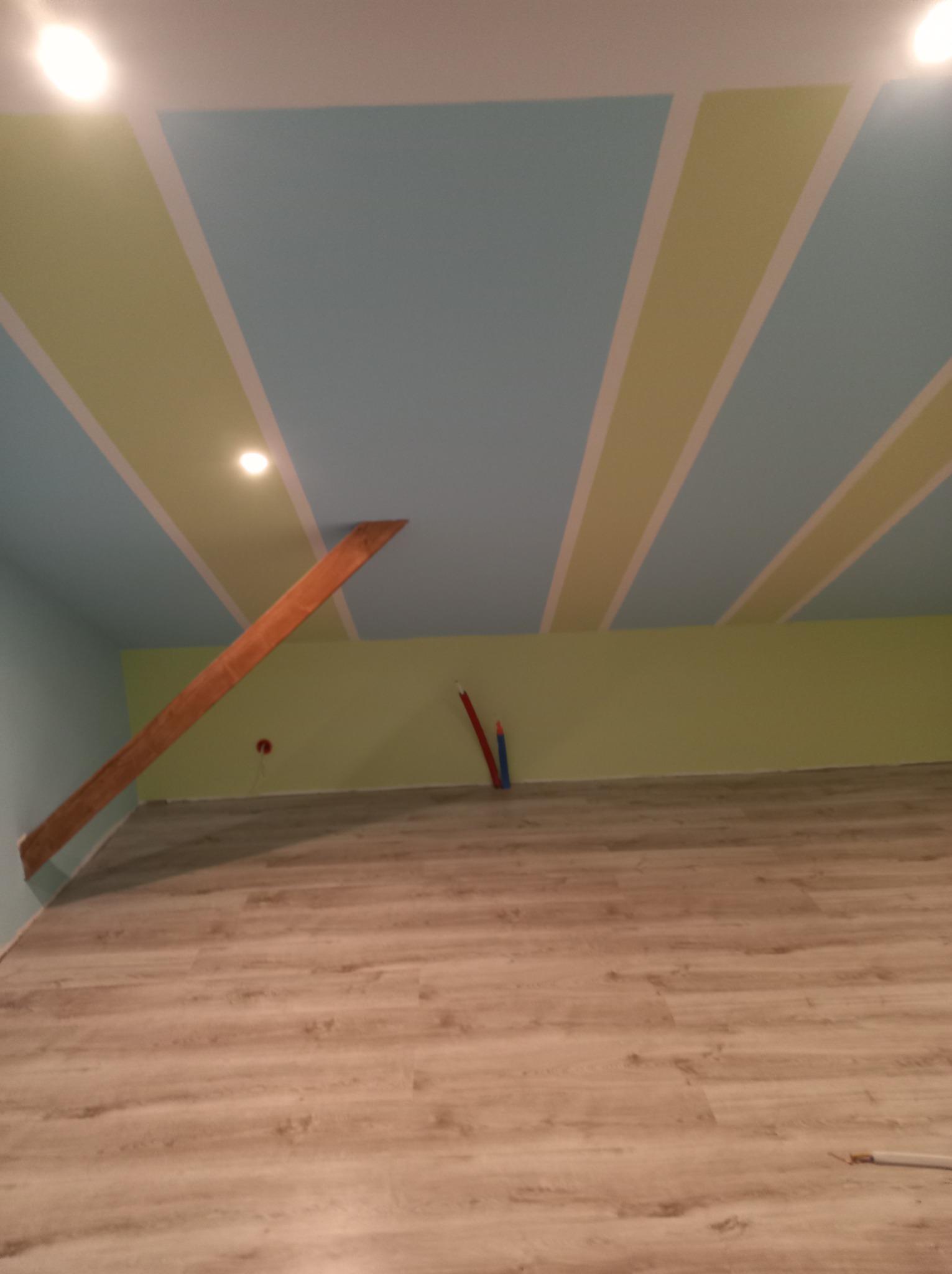 Pomieszczenie po remoncie: wzorzysty sufit w błękitno-zielone pasy, drewniana belka pod sufitem, ściany pomalowane na zielono, podłoga wyłożona panelami imitującymi drewno.