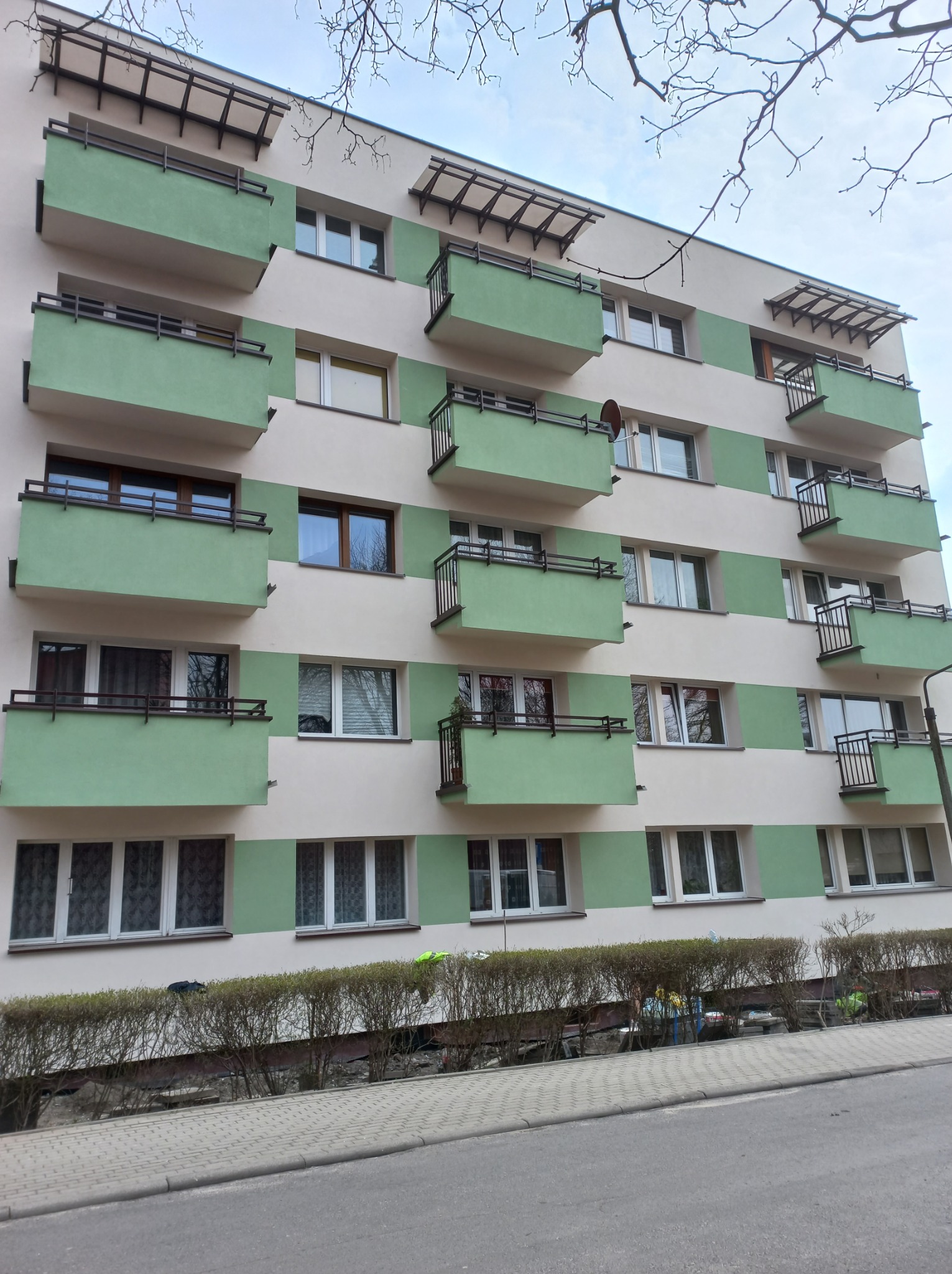 Wielopiętrowy budynek mieszkalny z zielonymi balkonami i elementami elewacji, widok z dołu, widoczne drzewa bez liści na tle nieba.