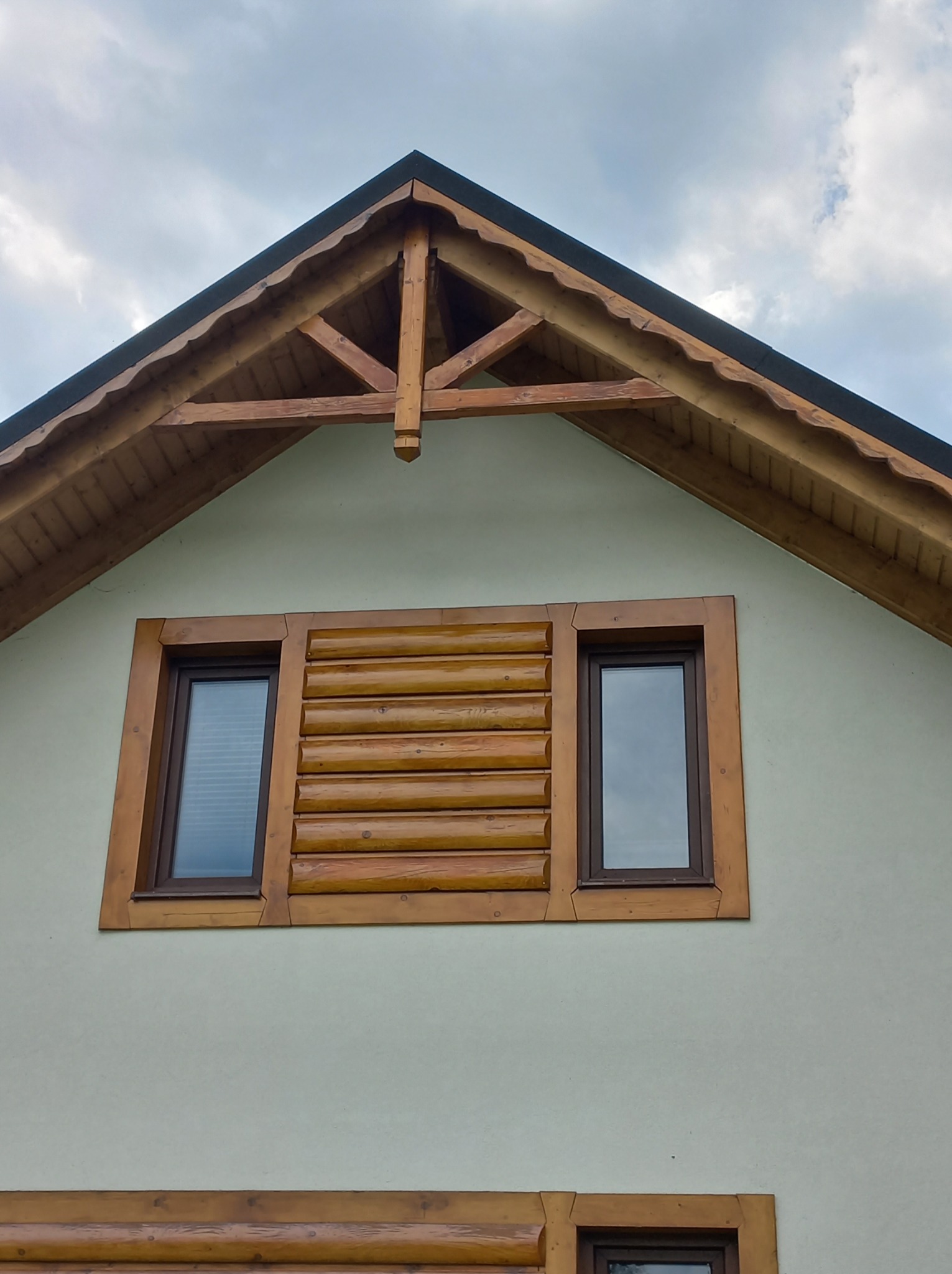 Szczyt domu z oknami w drewnianych ramach imitujących bale, drewniane elementy dekoracyjne pod dachem, jasna elewacja.