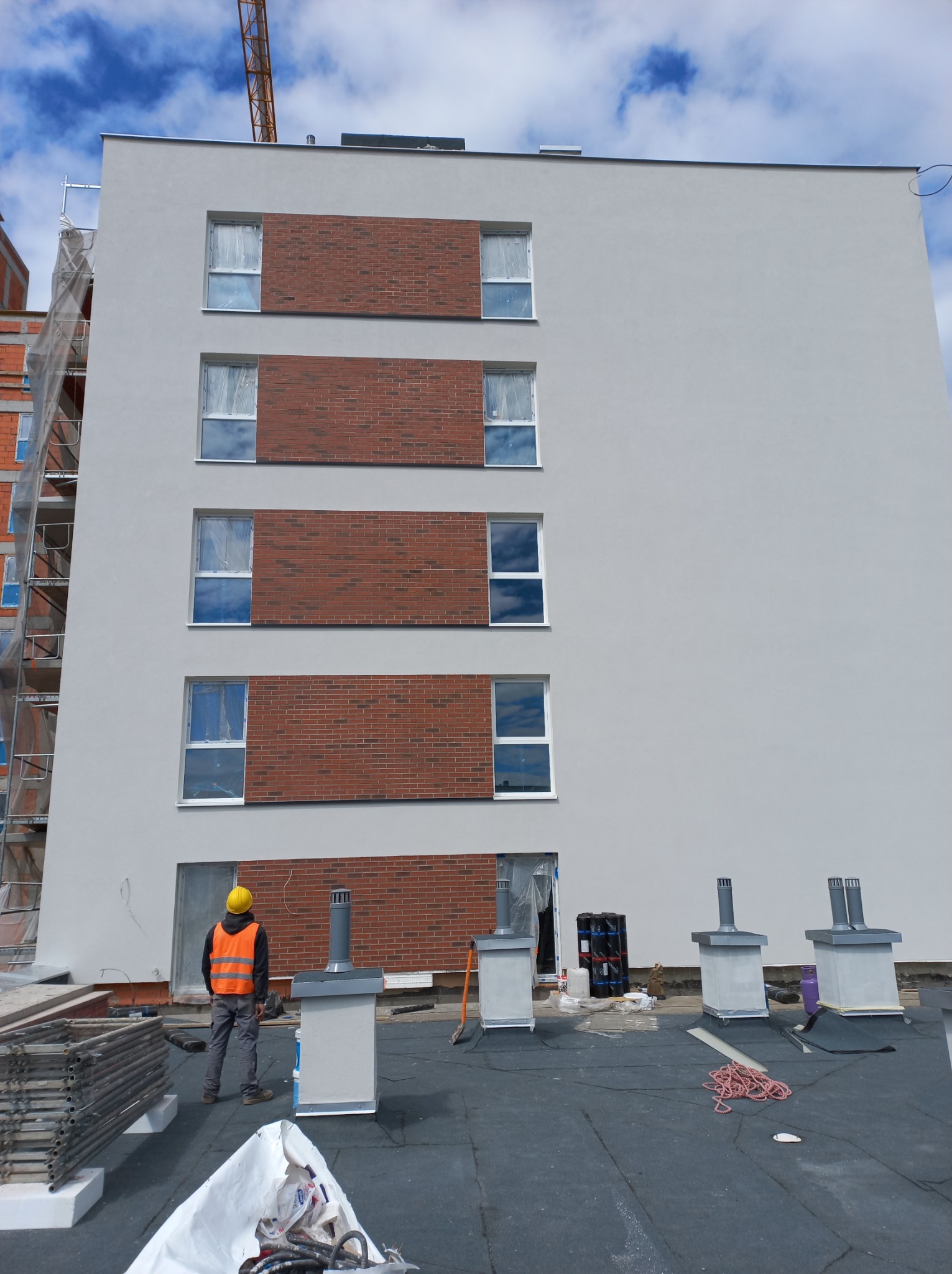 Budynek wielorodzinny z elewacją w trakcie wykańczania, widoczne fragmenty ceglanej okładziny wokół okien, robotnik w żółtym kasku i kamizelce odblaskowej na dachu, kominy wentylacyjne, rusztowania...
