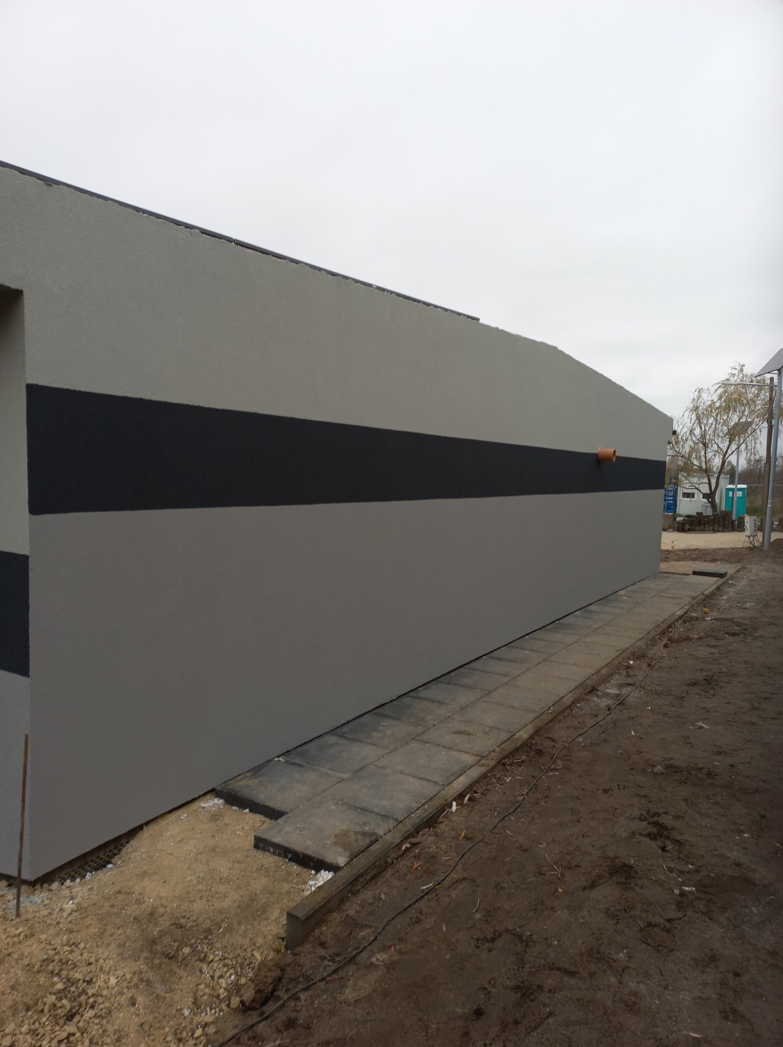 Szara elewacja budynku z ciemnoszarym pasem, widoczna rura spustowa i ułożona kostka brukowa wzdłuż ściany.