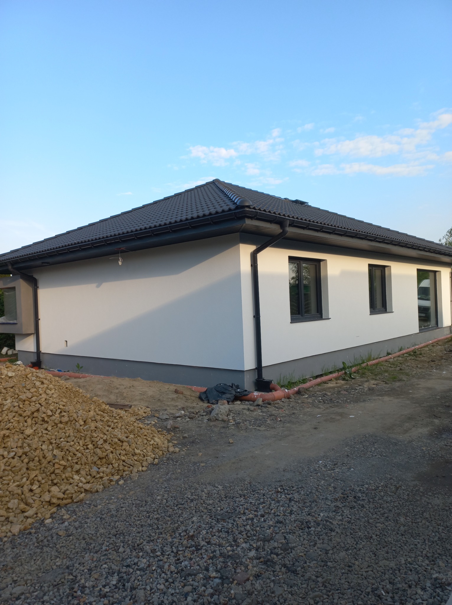 Jednopiętrowy dom z białą elewacją i ciemną dachówką, widoczny stos kamieni i rury drenażowe w trakcie prac wykończeniowych.