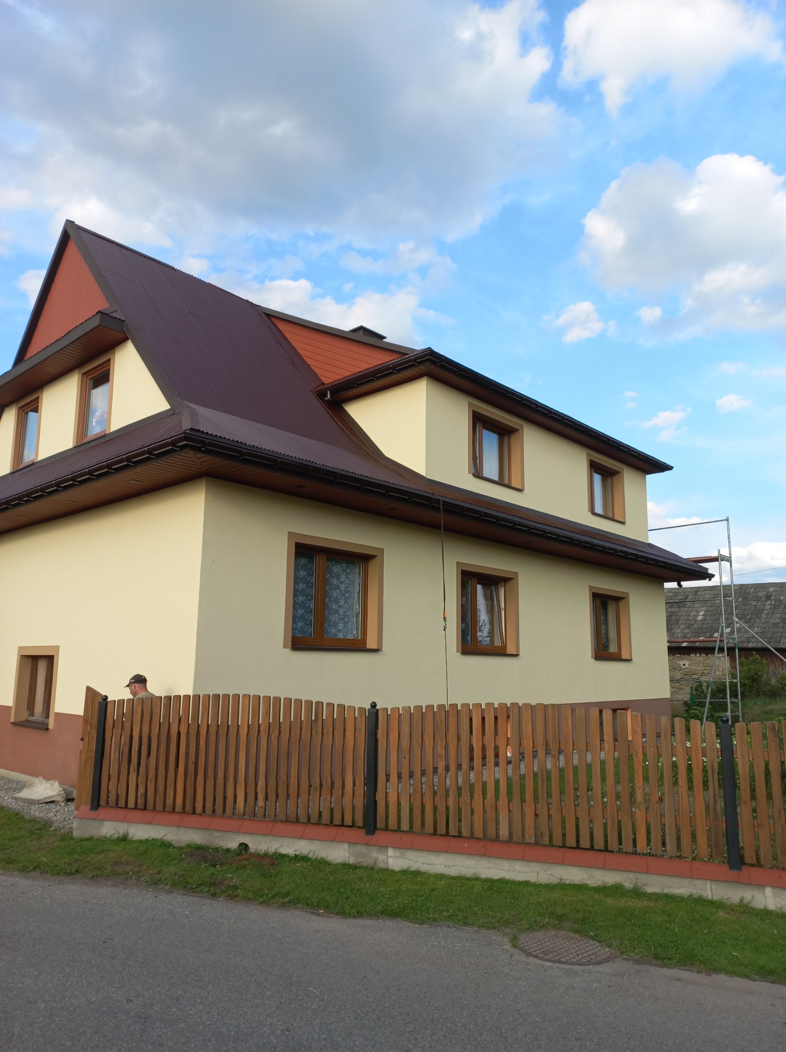 Dwupiętrowy dom z beżową elewacją i brązowym dachem, widziany z poziomu ulicy, z fragmentem drewnianego ogrodzenia w pierwszym planie i rusztowaniem z boku budynku.
