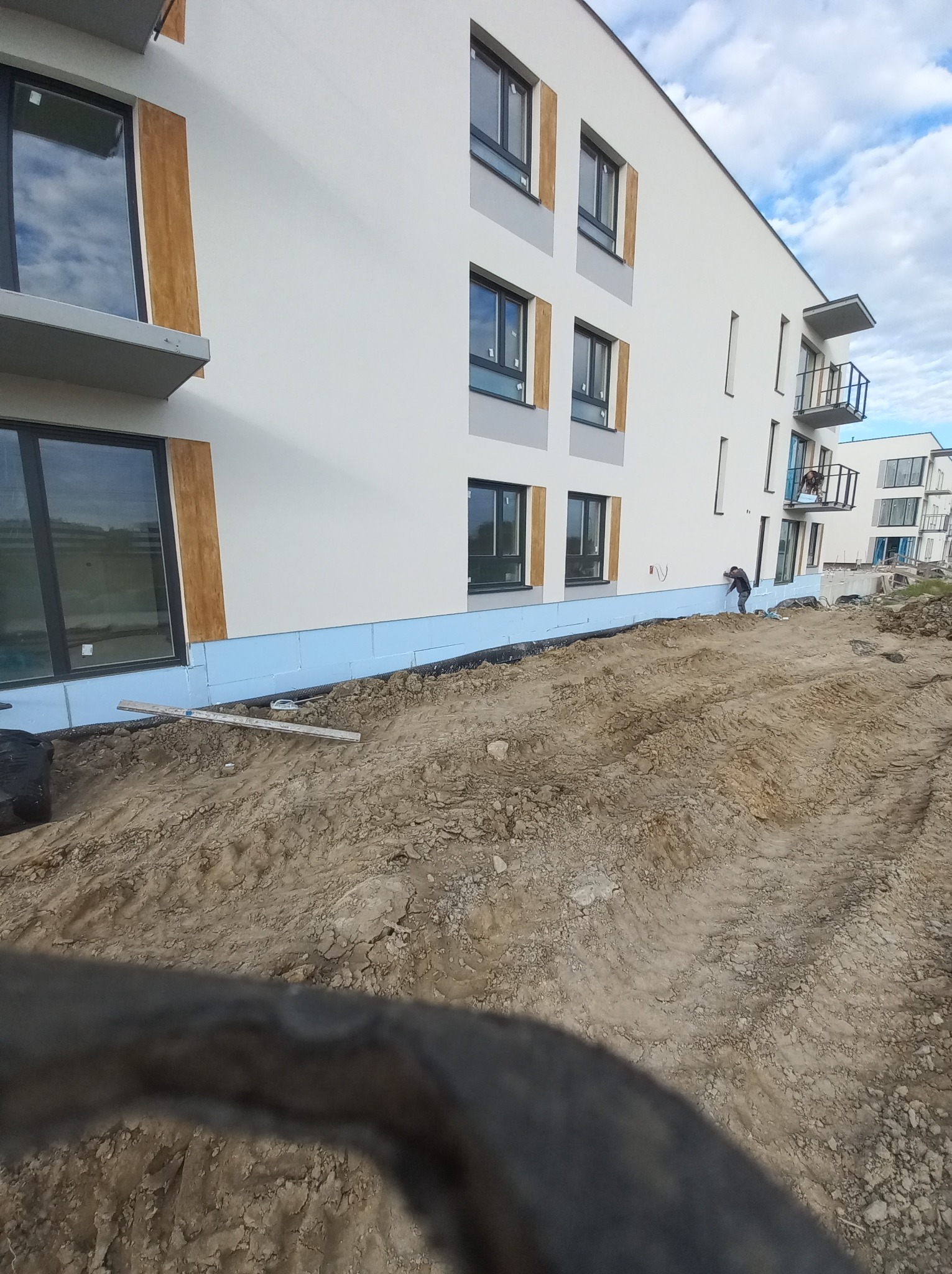 Prace przygotowawcze do ocieplenia elewacji budynku wielorodzinnego, widoczna warstwa izolacji przy gruncie i robotnik przy elewacji.