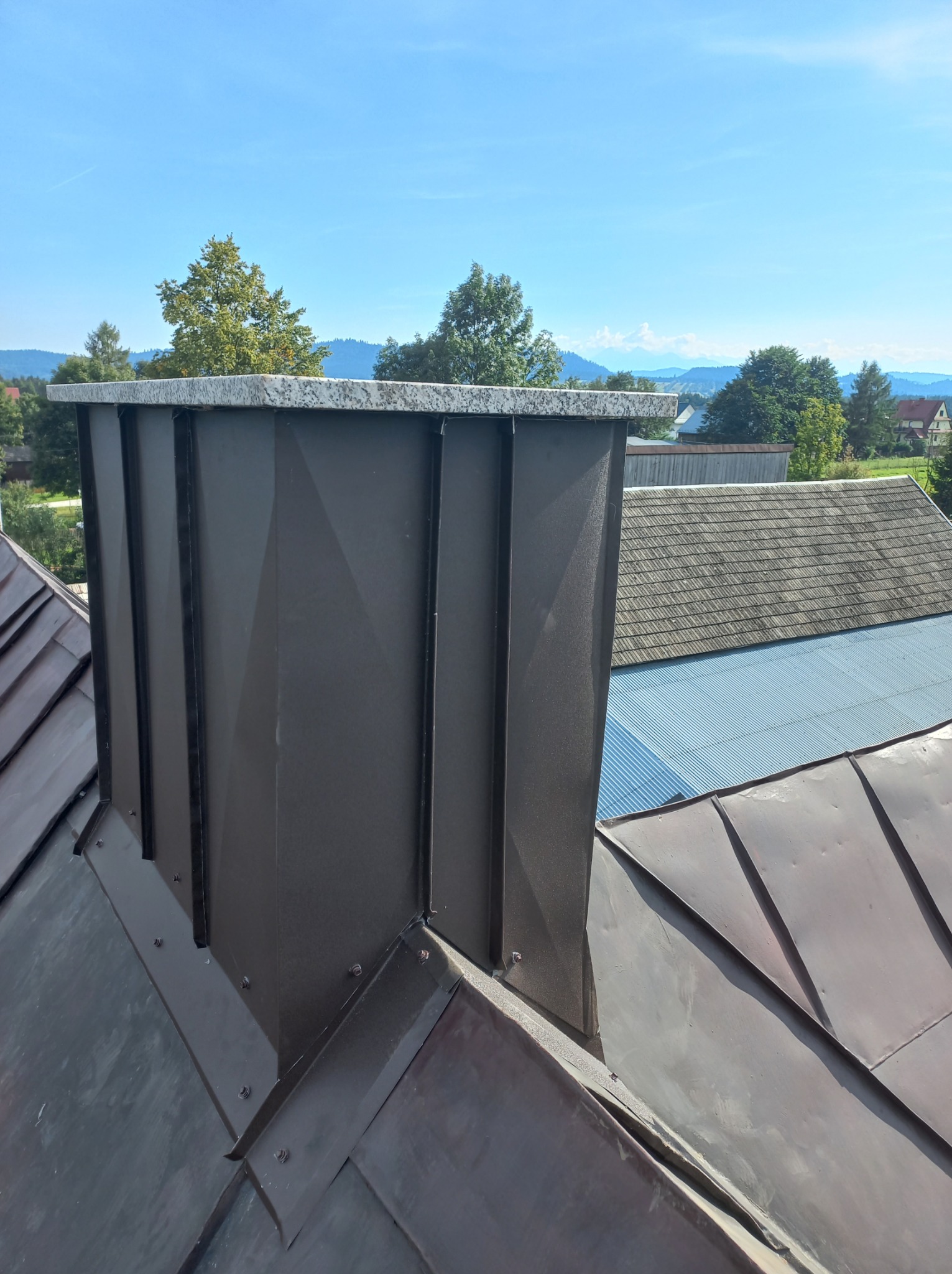 Nowoczesny, ciemny komin z kamiennym wykończeniem na dachu z metalowymi płytkami, w tle widok na zalesione wzgórza pod błękitnym niebem.