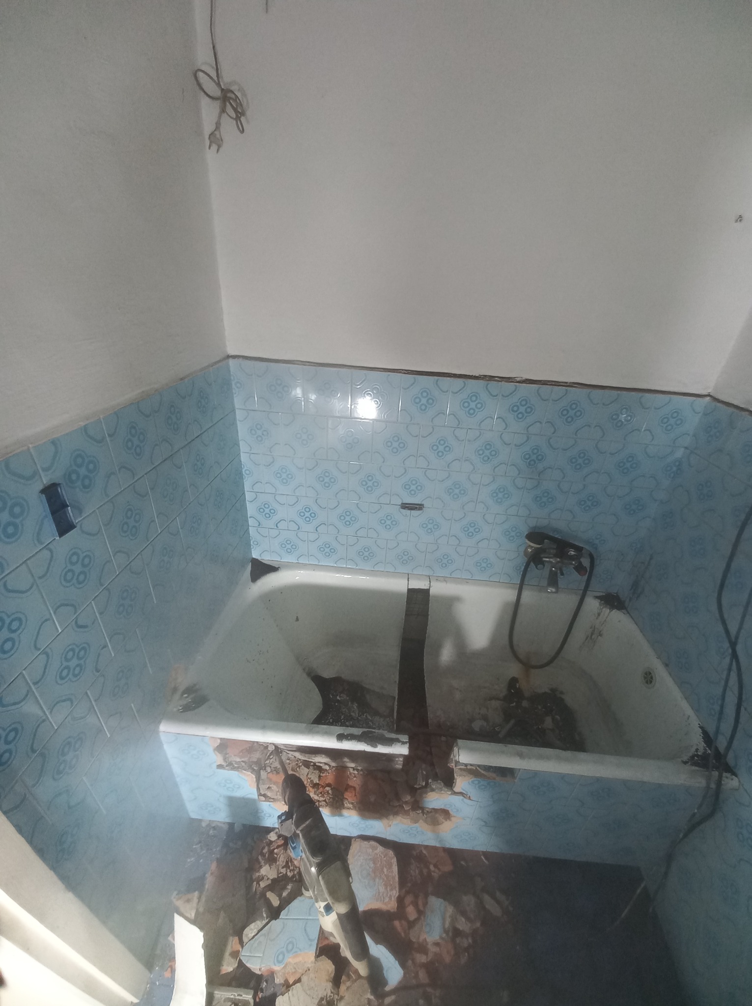 Demontaż starej, niebieskiej glazury w łazience za pomocą młota udarowego, widoczna wanna i fragment instalacji wodnej.