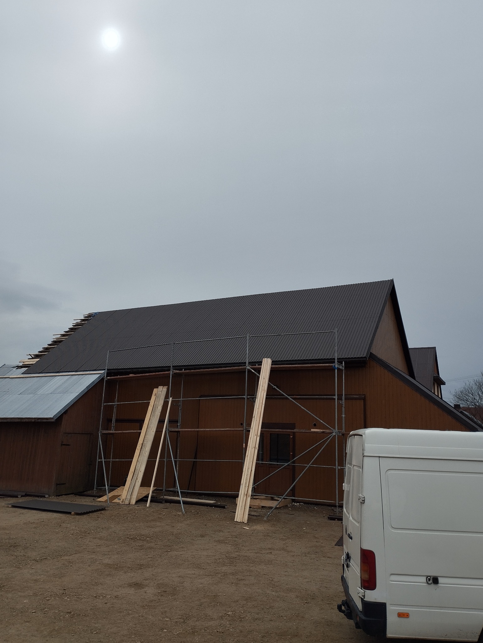 Montaż blachodachówki na drewnianym budynku, widoczne rusztowanie i deski przygotowane do montażu, pochmurne niebo.