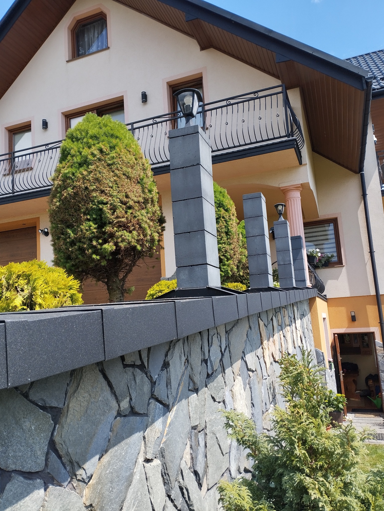 Nowoczesny kamienny mur z szarymi słupkami oświetleniowymi, otoczony zielenią, na tle dwupiętrowego domu z balkonem.