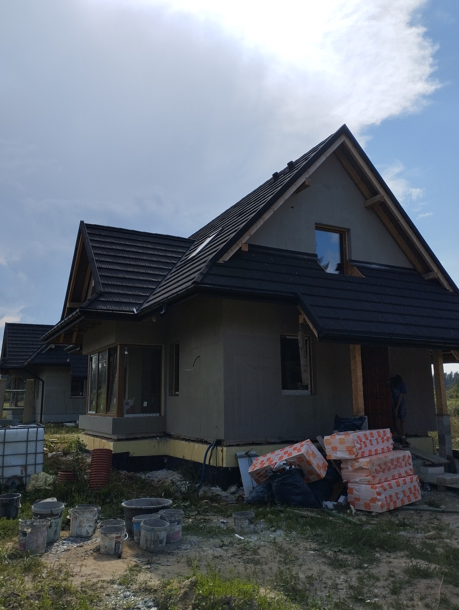 Dom w Frydmanie w trakcie ocieplania elewacji, widoczne materiały izolacyjne i puste wiadra po farbie, częściowo otynkowana fasada i czarna dachówka.