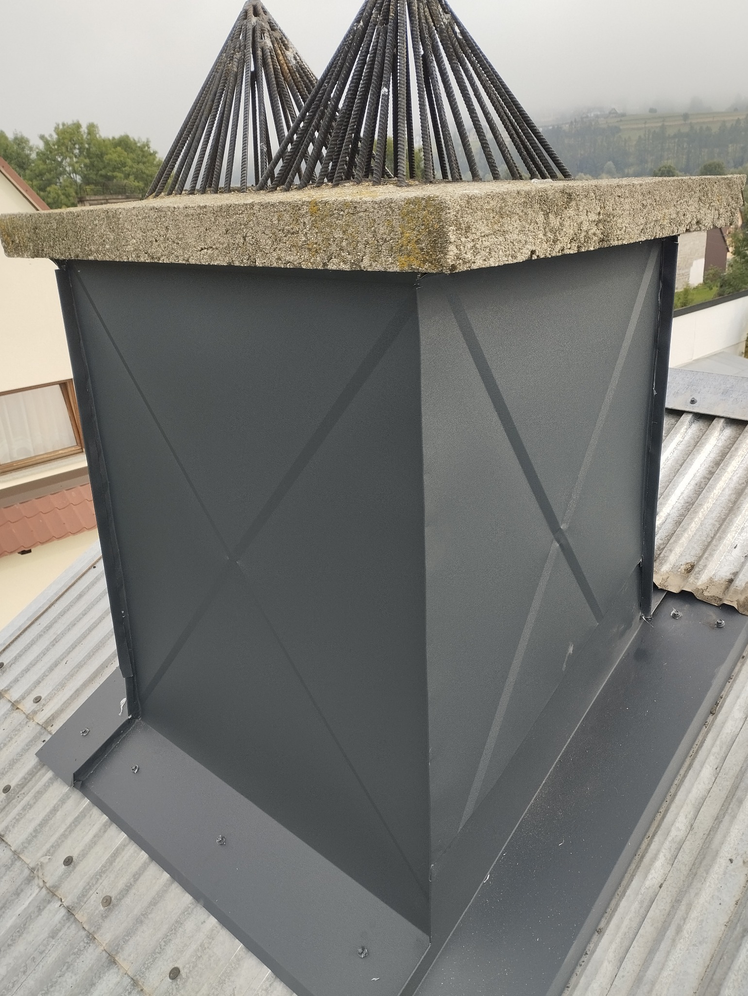 Nowoczesna obudowa komina z szarej blachy z ozdobnymi prętami na szczycie, zamontowana na dachu z blachy falistej, widok z bliska.