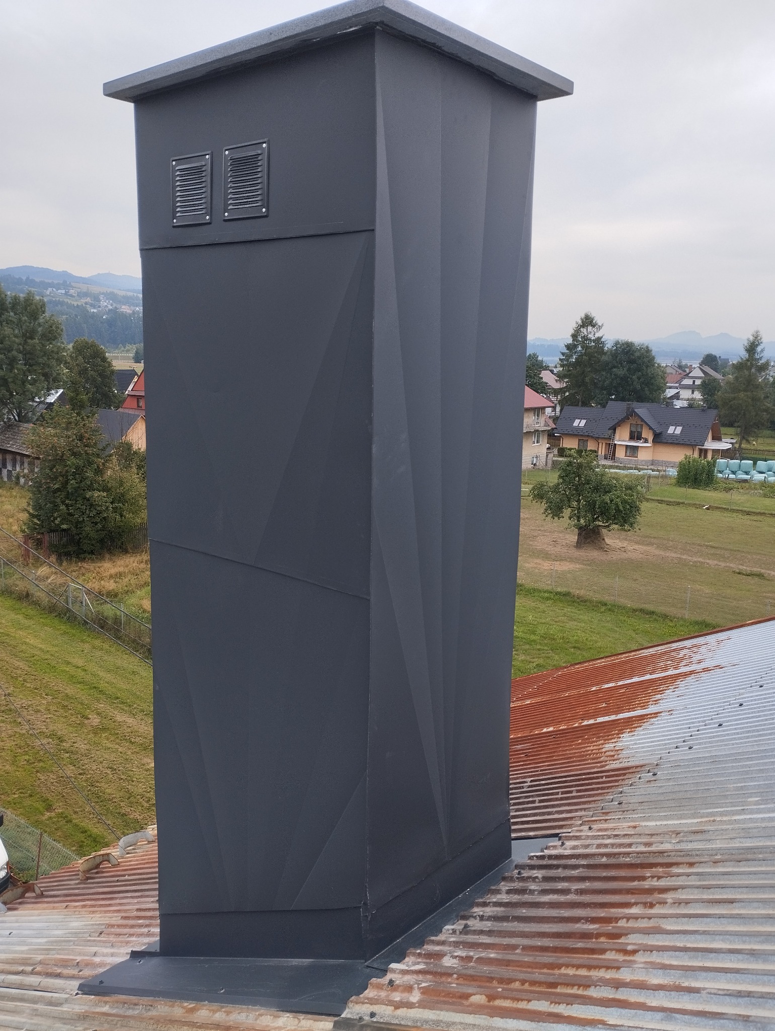 Szary, metalowy komin z geometrycznym wzorem na dachu z blachy trapezowej, w tle krajobraz wiejski z domami i wzgórzami.