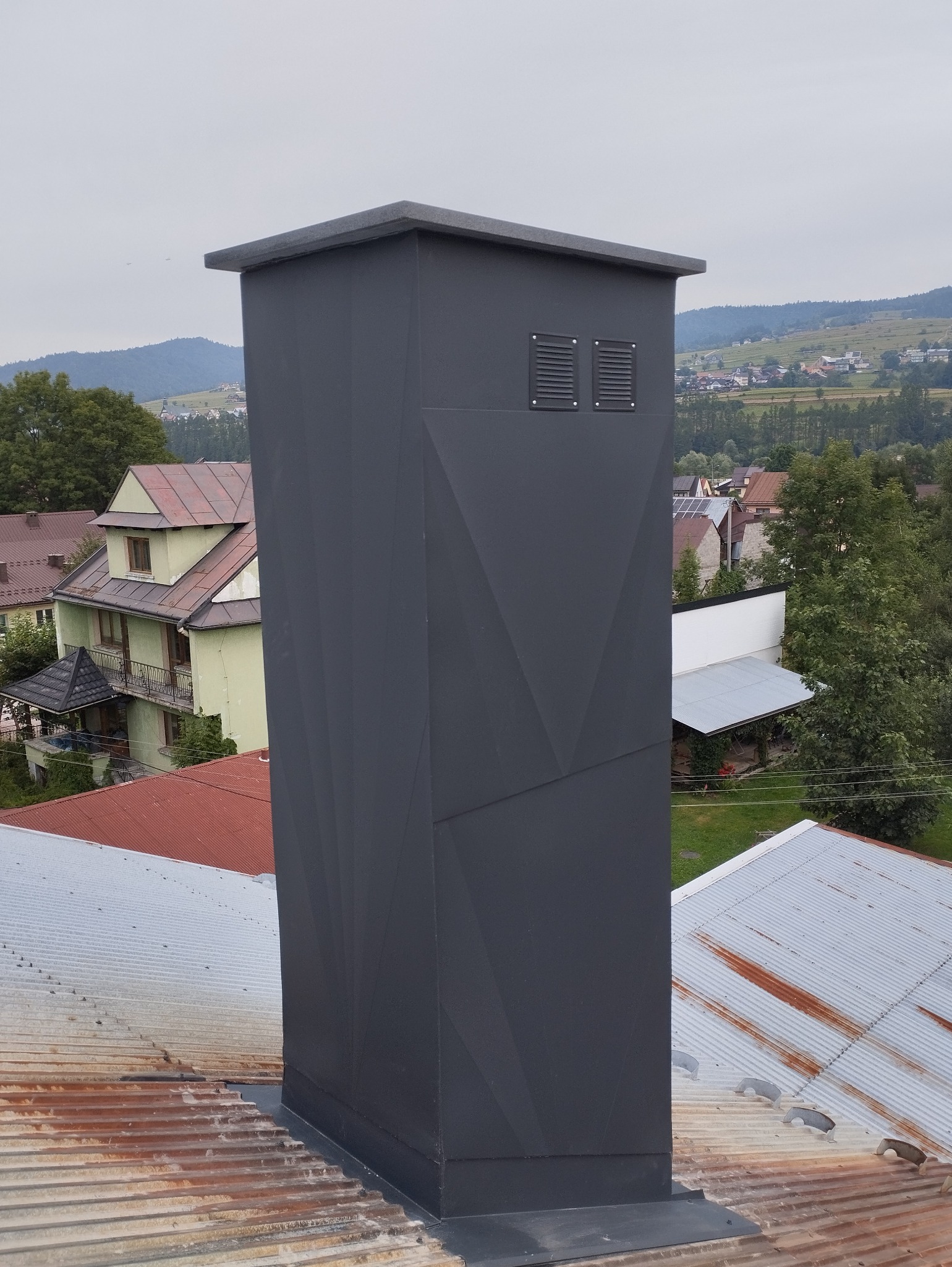 Nowoczesny, grafitowy komin z geometrycznym wzorem na dachu z blachy falistej, w tle zabudowa wiejska i zalesione wzgórza.