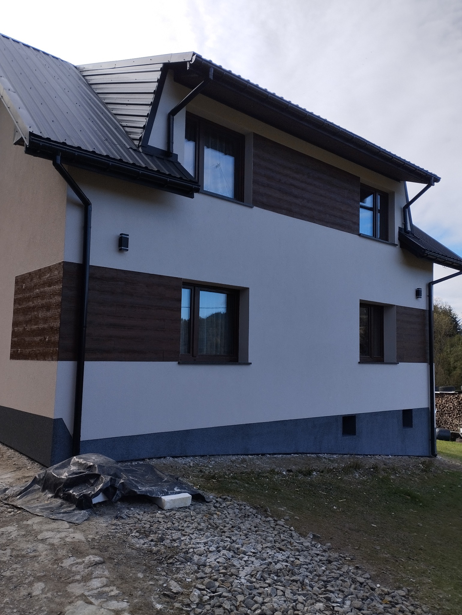 Dwukondygnacyjny dom z szarą elewacją i poziomymi panelami imitującymi drewno, ciemna dachówka i okna, widoczne przygotowania terenu wokół budynku.