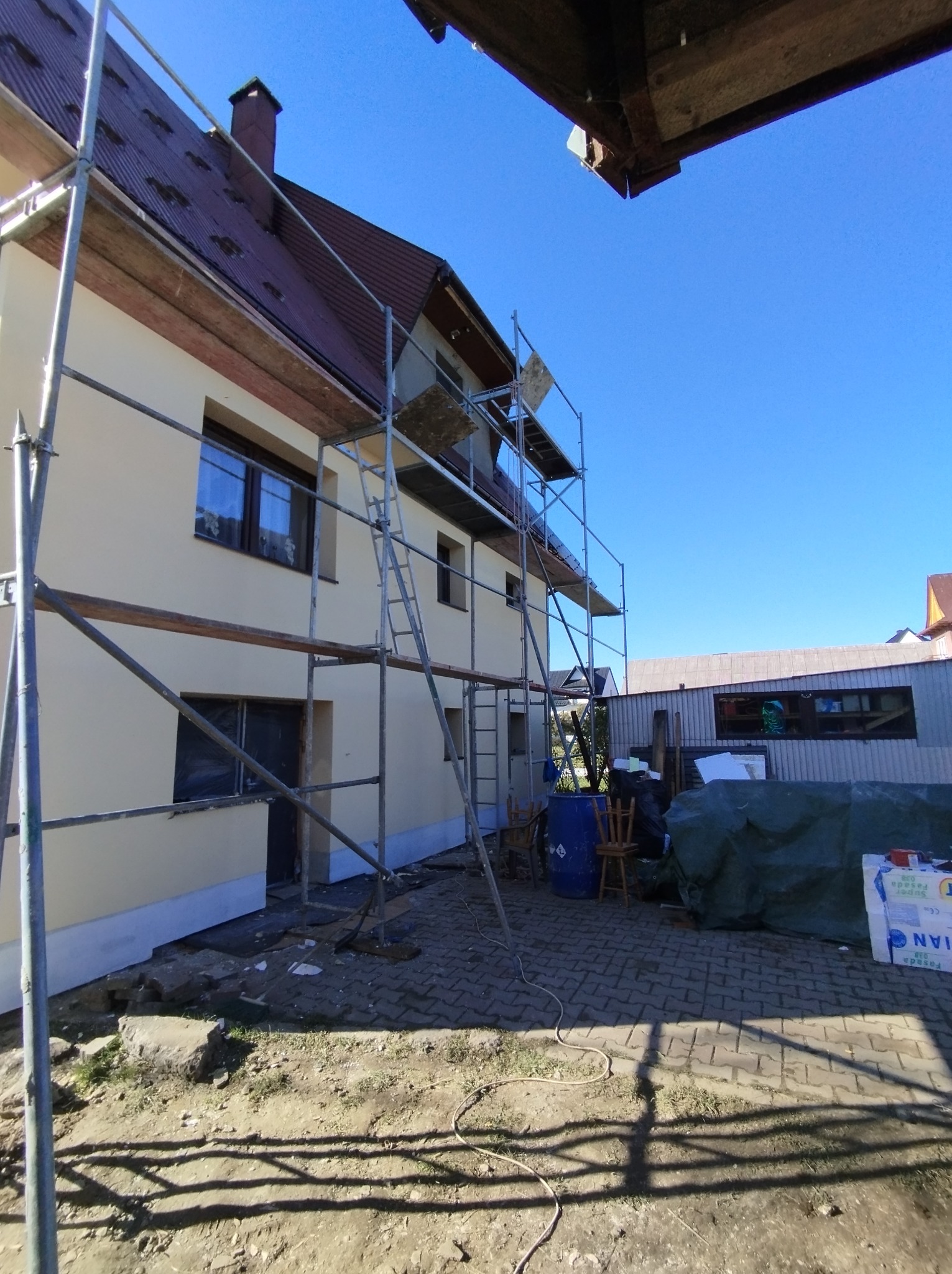 Dom jednorodzinny w trakcie prac elewacyjnych, widoczne rusztowania, fragment ocieplonej i pomalowanej ściany, zabezpieczone okna folią.