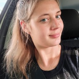 Julita Wisniewska - Portret uśmiechniętej blondynki z niebieskimi oczami w samochodzie, zapiętej pasami. Widoczna złota biżuteria i czarna błyszcząca koszulka.