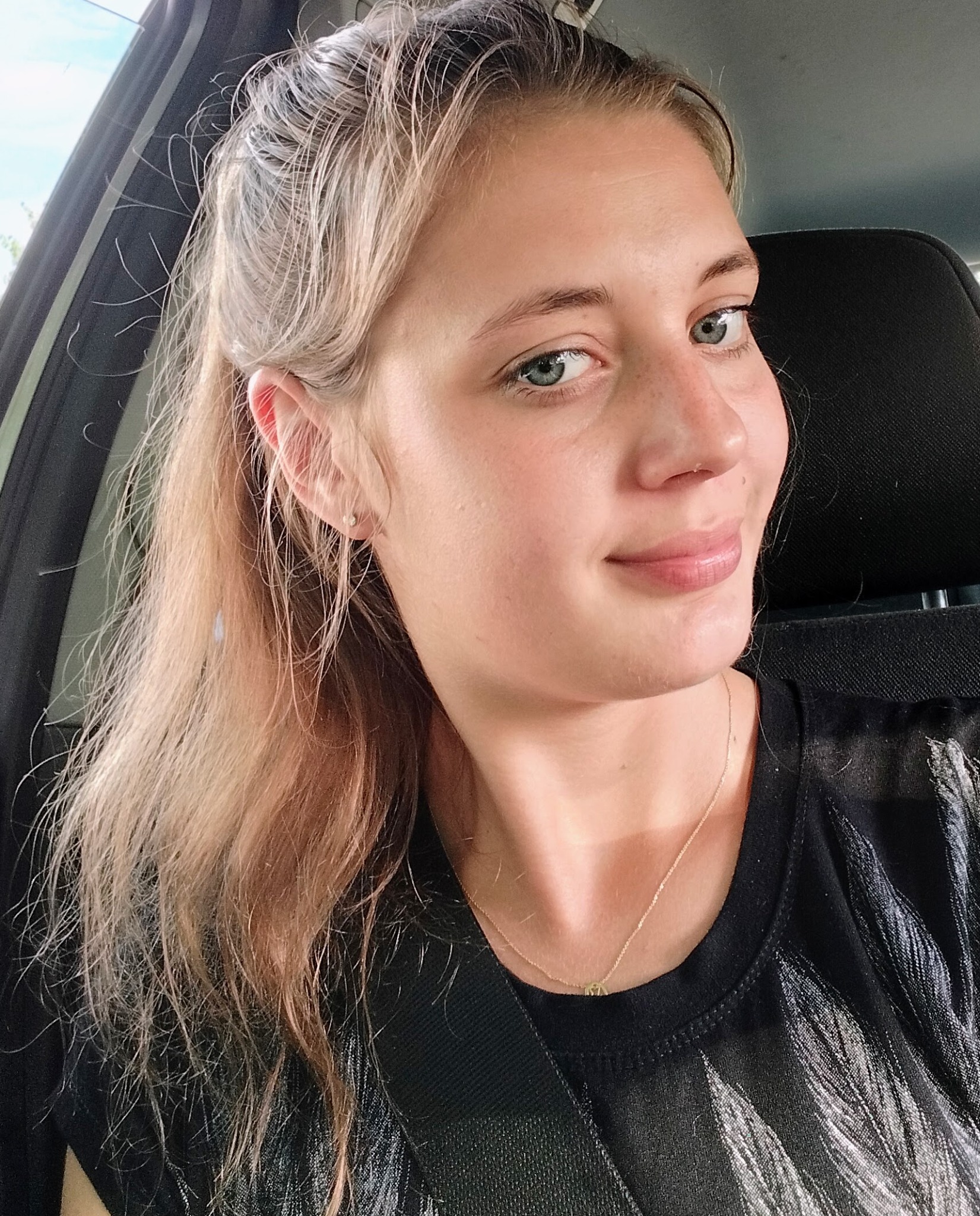 Portret uśmiechniętej blondynki z niebieskimi oczami w samochodzie, zapiętej pasami. Widoczna złota biżuteria i czarna błyszcząca koszulka.