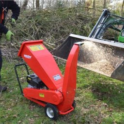 Lidia Derda pielęgnacja ogrodów - Czerwony rębak do gałęzi Hecht 6260 w trakcie pracy, wyrzucający zrębki do łyżki ładowarki, z stertą gałęzi w tle, podczas wycinki drzew.