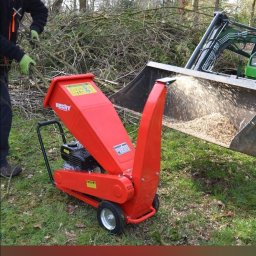 Lidia Derda pielęgnacja ogrodów - Czerwony rębako-rozdrabniacz gałęzi marki Hecht w trakcie pracy, z boku widoczny podnośnik z załadunkiem zrębków, w tle sterta gałęzi.