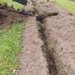 Lidia Derda pielęgnacja ogrodów - Wykonanie wykopu wąskorowowego w trawniku za pomocą mini-koparki łańcuchowej, widoczny świeżo wykopany rów z wystającą trawą, w tle ogrodzenie i elementy architektury ogrodowej.