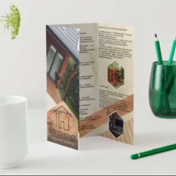 Trójdzielna ulotka reklamowa firmy Woodhouse z wizualizacjami domów, stojąca obok białego kubka, wazonu z zielonymi roślinami i zielonego pojemnika z kredkami na białym tle.