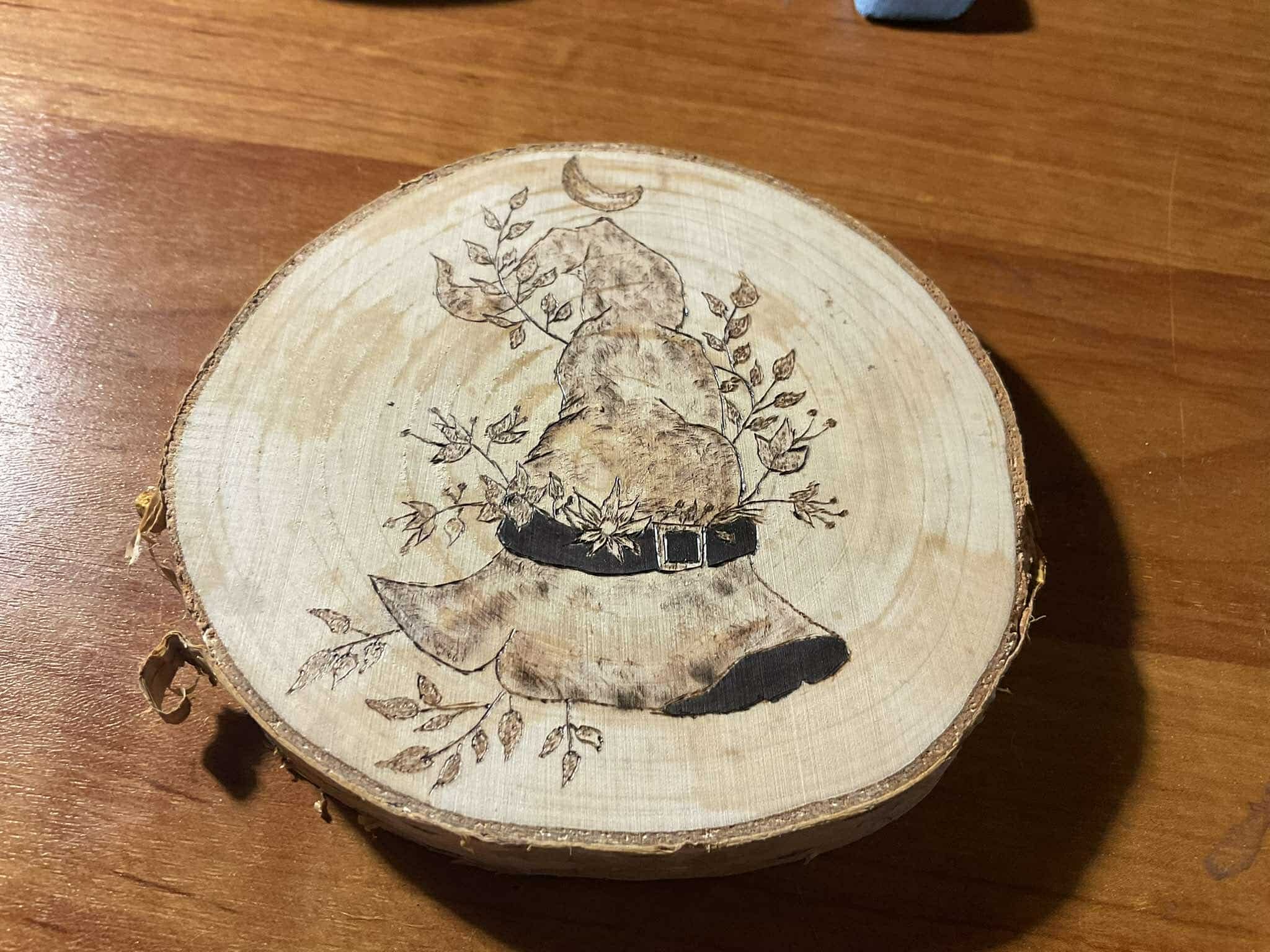 Ręcznie wypalana drewniana podkładka pod kubek z motywem czapki czarownicy otoczonej liśćmi i półksiężycem, widoczna struktura drewna i kora na obrzeżach.