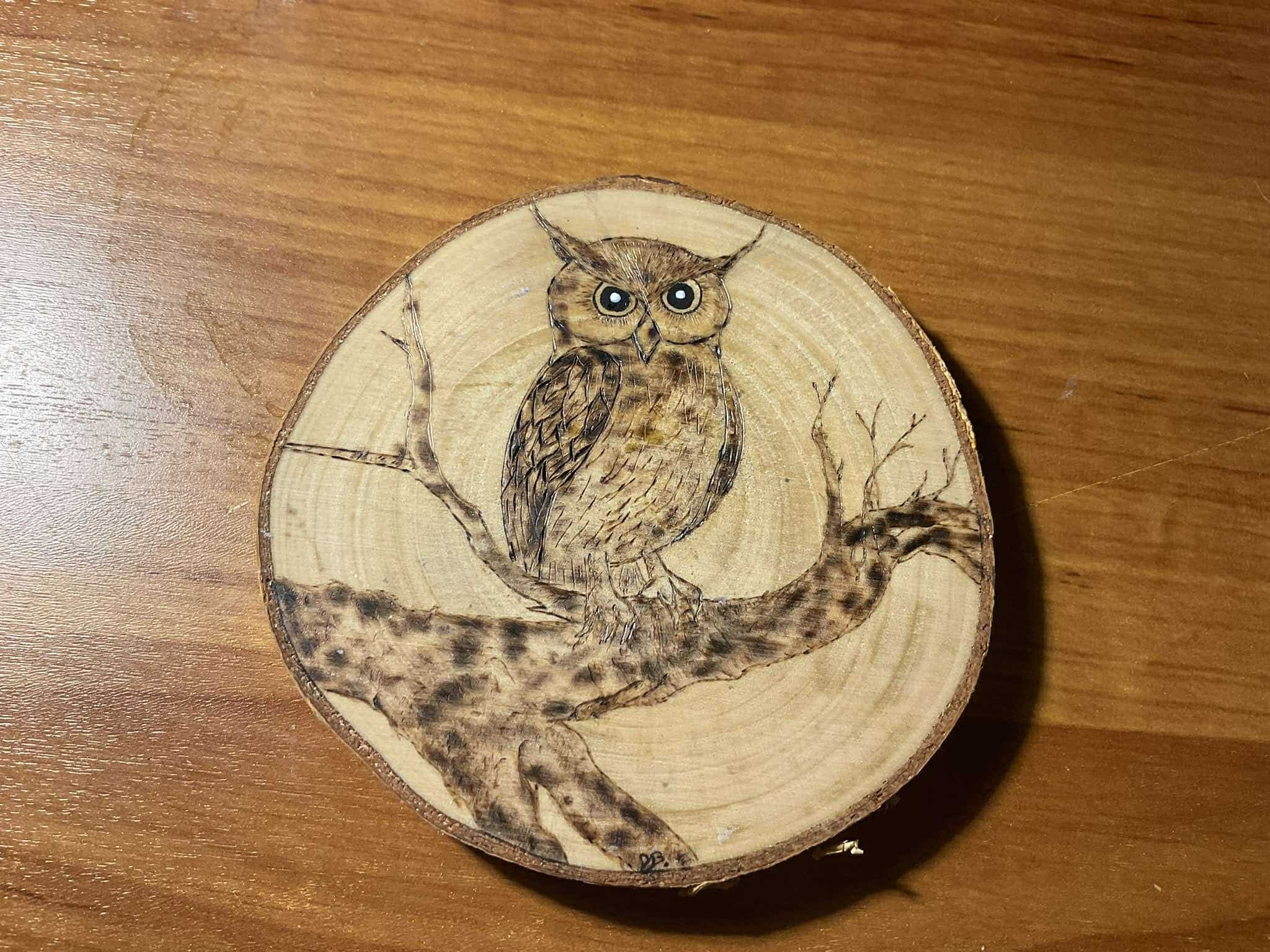 Ręcznie wypalana, drewniana podkładka pod kubek z wizerunkiem sowy siedzącej na gałęzi, umieszczona na drewnianym stole.