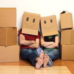 Błyskawiczne przeprowadzki - Para siedzi na podłodze z kartonami, mają na głowach kartony z narysowanymi uśmiechniętymi buziami, skrzyżowane ręce, bose stopy.