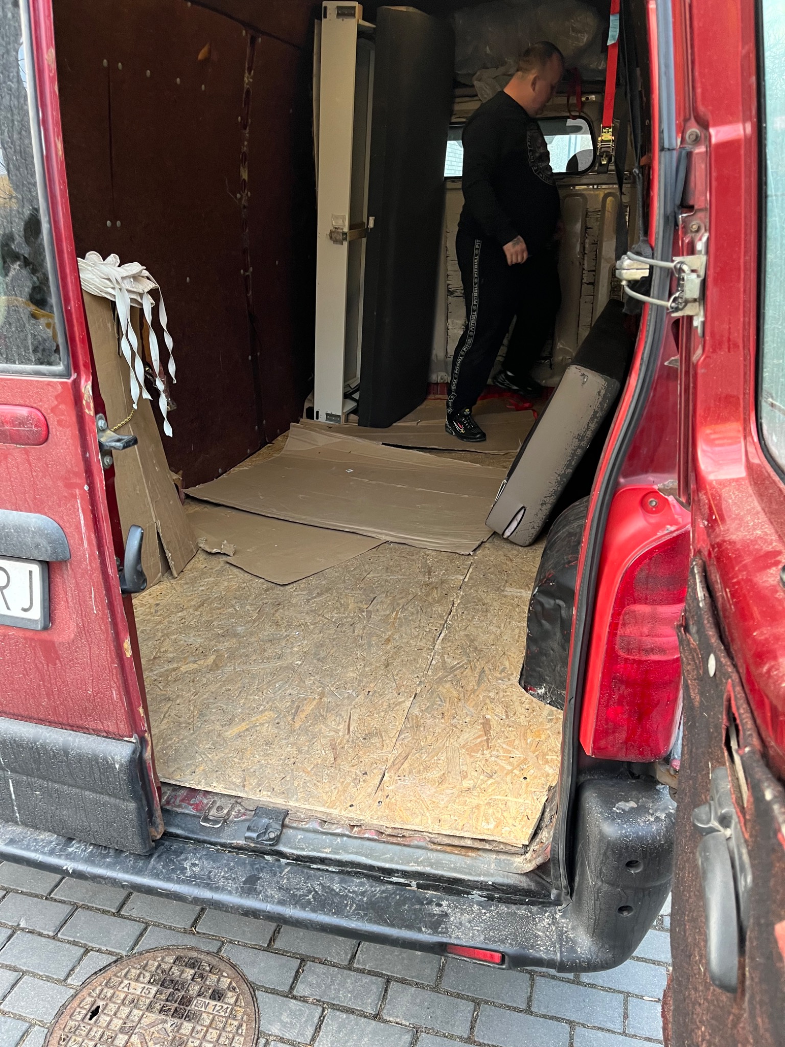 Wnętrze czerwonego busa z otwartymi drzwiami bocznymi, częściowo załadowane meblami i kartonami, widoczna sylwetka mężczyzny w czarnym ubraniu.