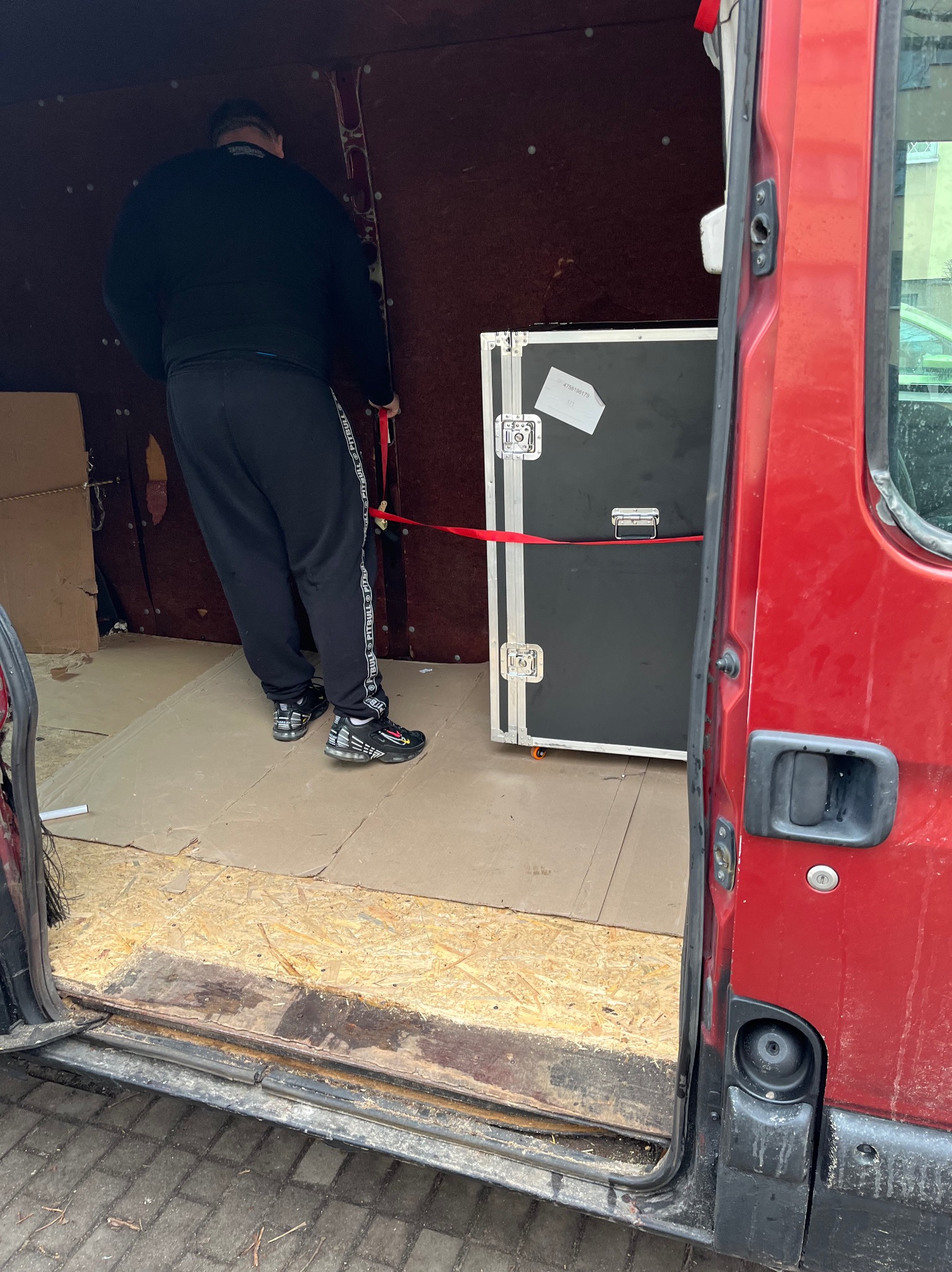 Mężczyzna w czarnym dresie zabezpiecza skrzynię transportową czerwoną taśmą wewnątrz czerwonego busa z otwartymi drzwiami, widoczna podłoga wyłożona kartonem.