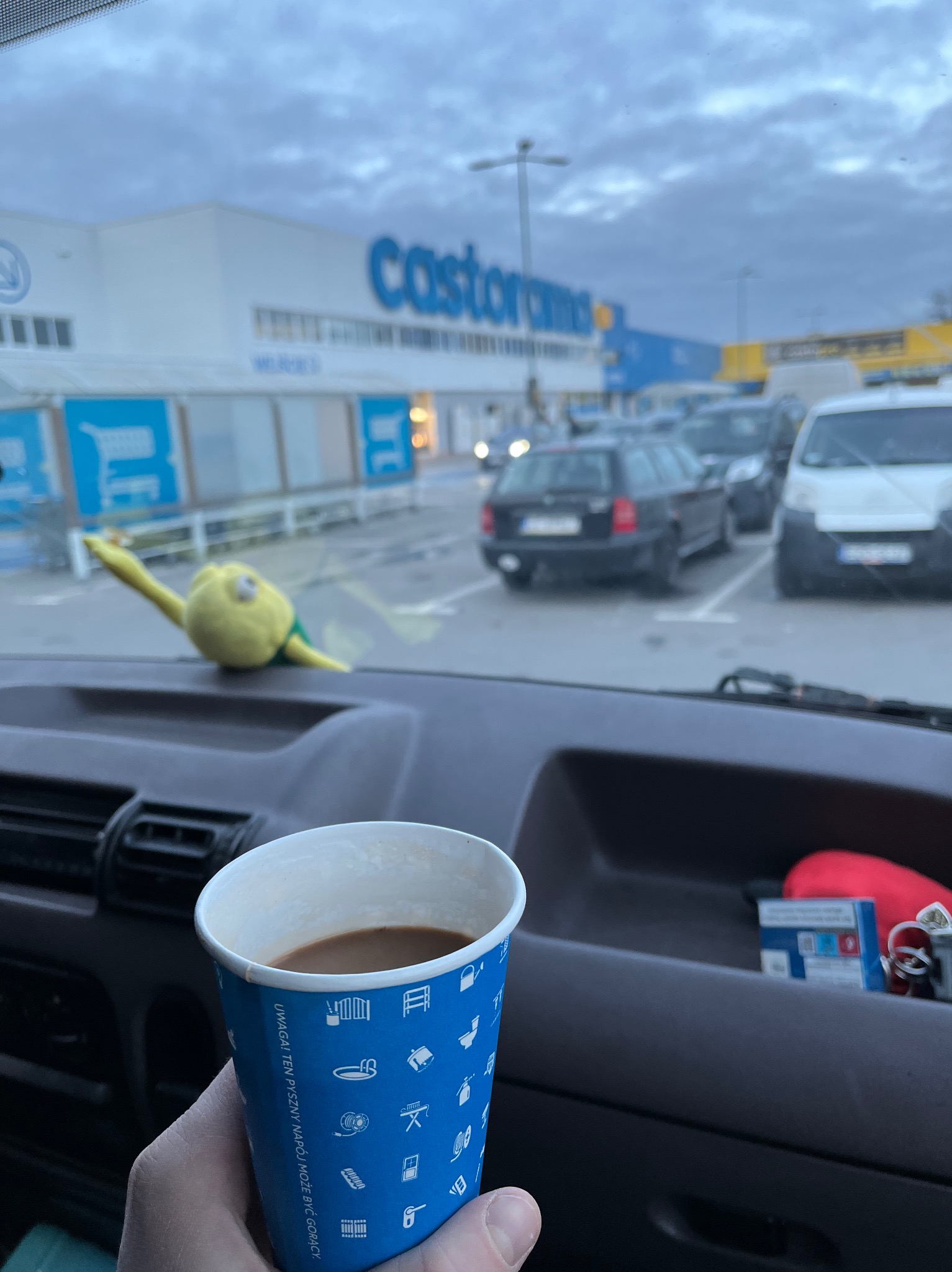 Kawa w papierowym kubku z niebieskimi ikonami, widziana z wnętrza samochodu dostawczego na parkingu przed sklepem Castorama, z żółtą maskotką na desce rozdzielczej.