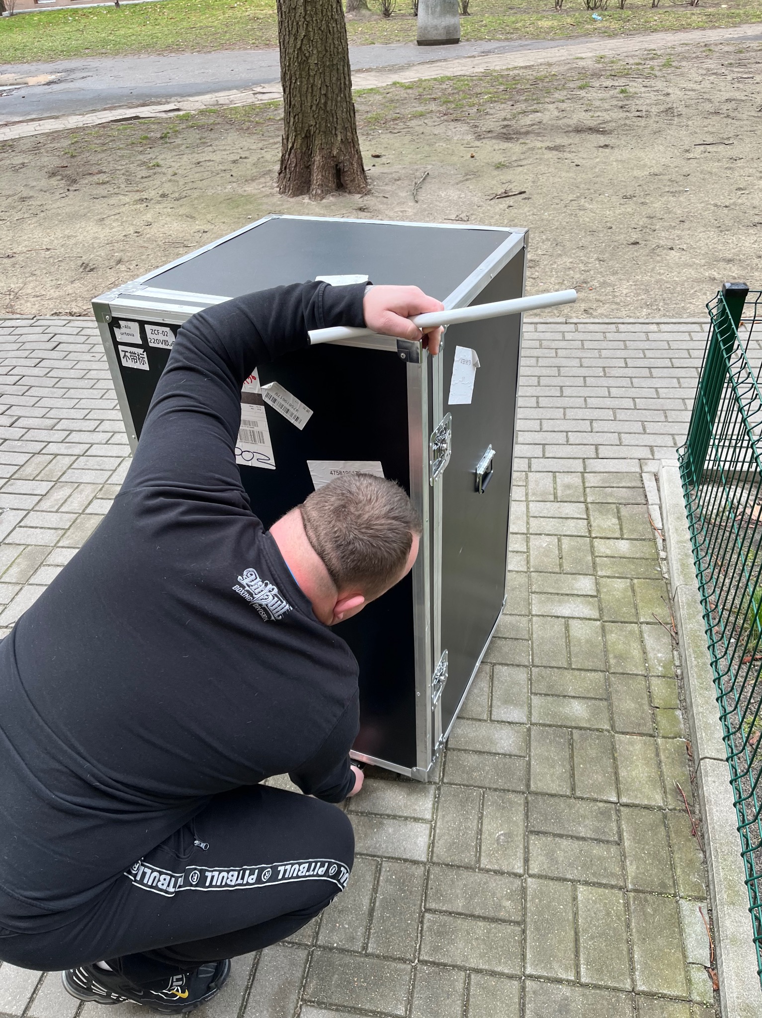 Mężczyzna w czarnym dresie z napisem 'PITBULL' montuje białą rurkę w czarną skrzynię transportową na chodniku z kostki brukowej, widok z tyłu.