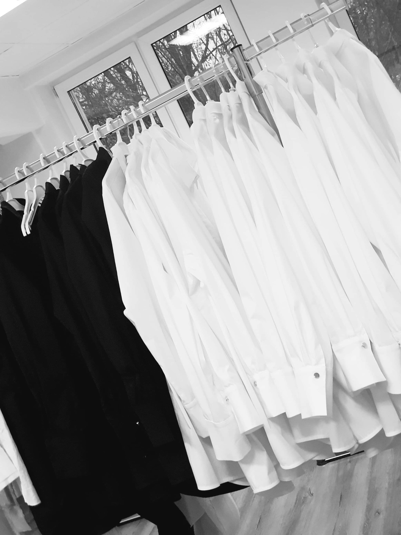 Rząd białych i czarnych koszul męskich na wieszakach, widok z dołu, monochromatyczne ujęcie z oknem w tle.